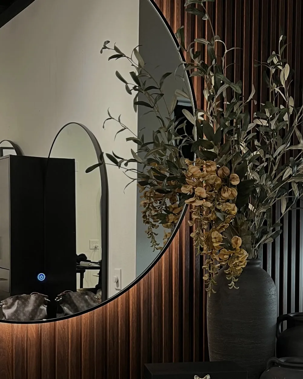 Inside Belleza De Oro Salon: a backlit round mirror on a dark wood slat wall with olive branches