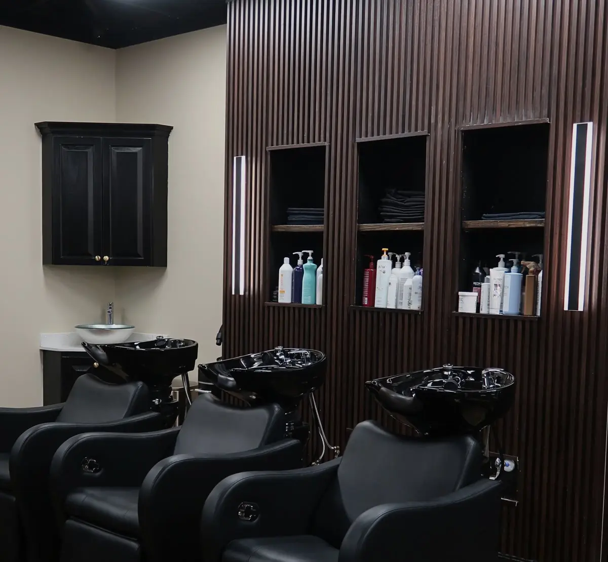 Three black shampoo bowls in front of a wood slat wall with lit product shelves